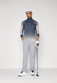 Herremote golfklær for menn, med en langermet topp i ombre blå og grå, lyse grå bukser, hvite sko og en hvit caps, som holder en golfkølle.