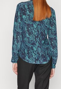 Blouse à manches longues en bleu foncé avec un motif tourbillonnant turquoise et noir. Présente un ourlet arrondi et des manches à poignets.