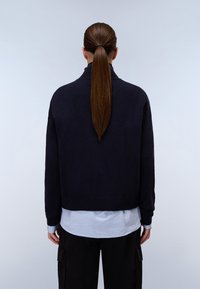 Marineblaues Strickpullover mit umgeschlagenem Kragen, cropped Passform und geripptem Saum, getragen über einem hellblauen Hemd. Die Rückansicht zeigt langes braunes Haar, das nach hinten gebunden ist.