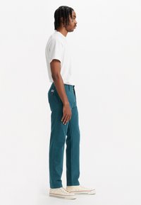 Pantaloni din bumbac teal cu o croială lejeră, buzunare standard și o marcă subtilă pe spate. Împreună cu un tricou alb simplu și adidași.