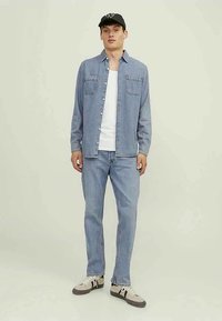 Camisa de ganga azul claro, top branco, jeans azul claro combinando e ténis bege. Boné de basebol preto com detalhe de logótipo.