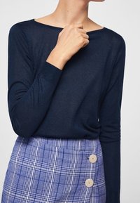 Osoba ubrana w granatową bluzkę z długim rękawem i niebieską spódnicę w kratę z dwoma dużymi beżowymi guzikami po bokach. Ręka blisko obojczyka.