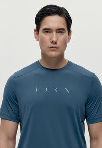 Muška atletska majica u tamnoplavoj boji, izrađena od glatkog, rastezljivog materijala. Ima kratke rukave i suptilan sivi logotip otisnut na prsima.