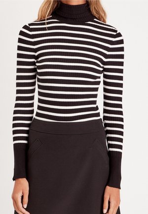 Femme portant un pull à col roulé côtelé rayé noir et blanc et une jupe noire taille haute avec des poches latérales.