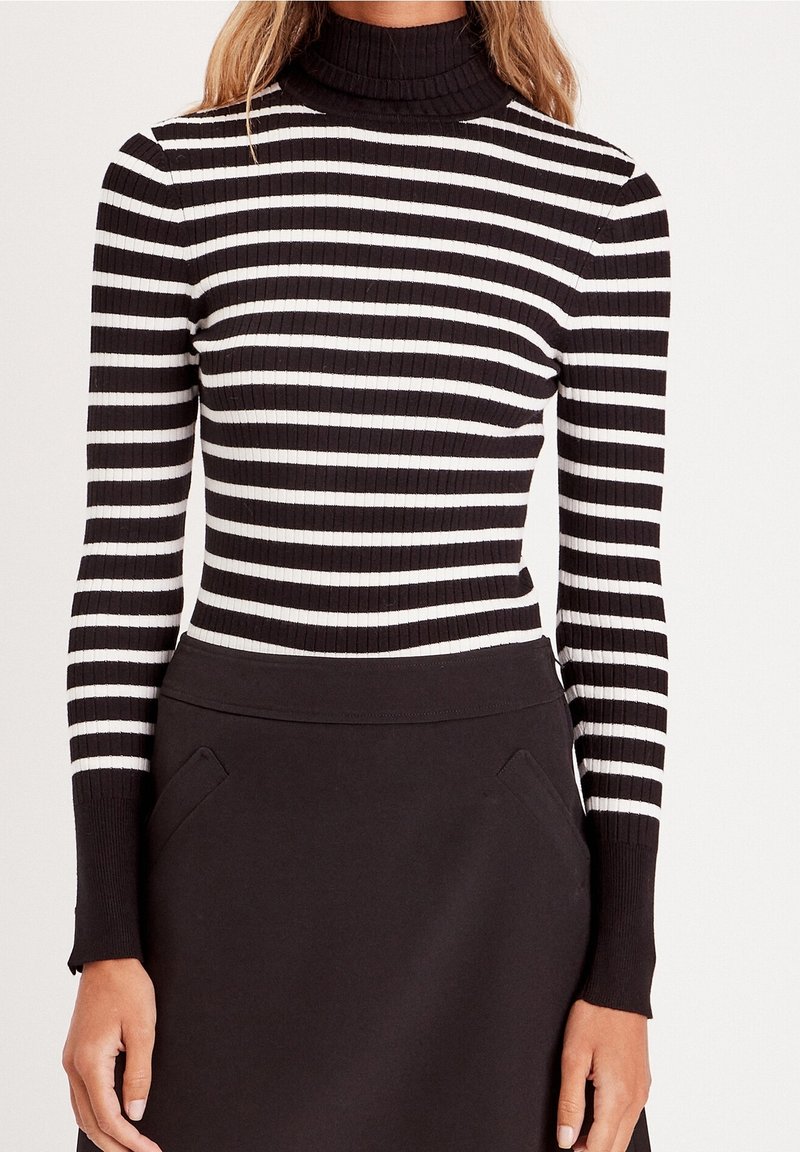 Femme portant un pull à col roulé côtelé rayé noir et blanc et une jupe noire taille haute avec des poches latérales.