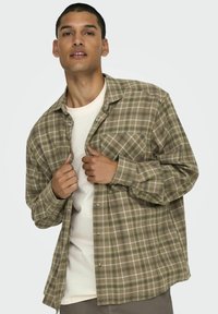 Koszula w kratę w odcieniach zieleni i brązu, z guzikami, kieszenią na piersi i długimi rękawami. Nosi się ją na jasnym T-shircie.