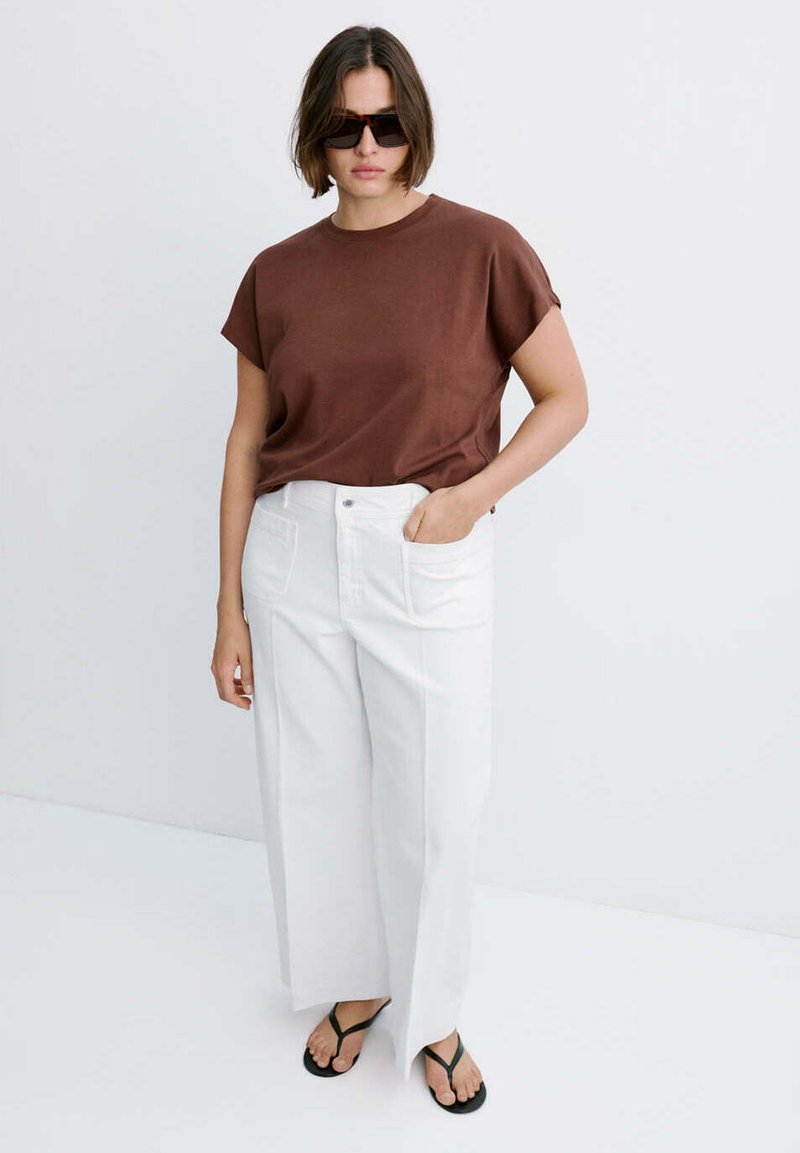 Femme portant un haut marron à manches courtes, un pantalon blanc à jambes larges, des tongs noires et des lunettes de soleil foncées, debout devant un fond clair uni.