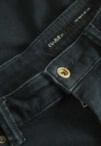 Tumedast sinised denim püksid, millel on metallik kuldne nupp ja must brändi silt, sujuva tekstuuriga ja klassikalise viie taskuga disain.