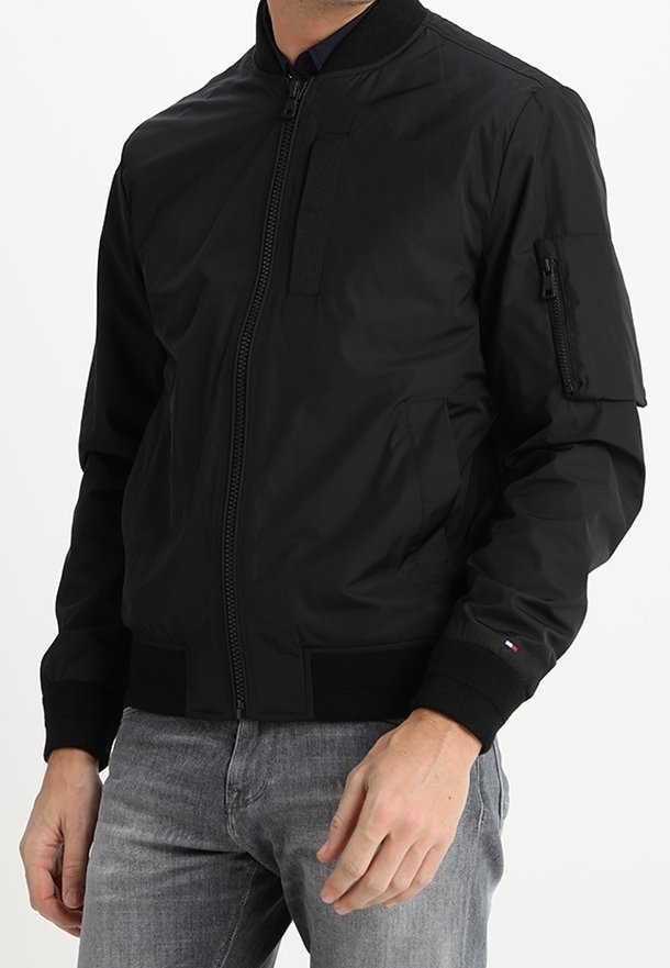 Veste bomber noire zippée avec col, poignets et ourlet côtelés, portée avec un jean gris, avec une poche zippée sur la partie supérieure gauche de la manche.