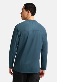 Maglietta a maniche lunghe di colore teal, tessuto testurizzato, design senza cuciture sulle spalle, scollatura tonda, indossata con pantaloni cargo neri, vista da dietro.