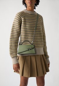 Πράσινη δερμάτινη τσάντα χειρός με δομημένο σχήμα, ασημένιο λογότυπο και αλυσίδα ως λουρί, συνδυασμένη με ριγωτό πλεκτό πουλόβερ και πλισέ φούστα.
