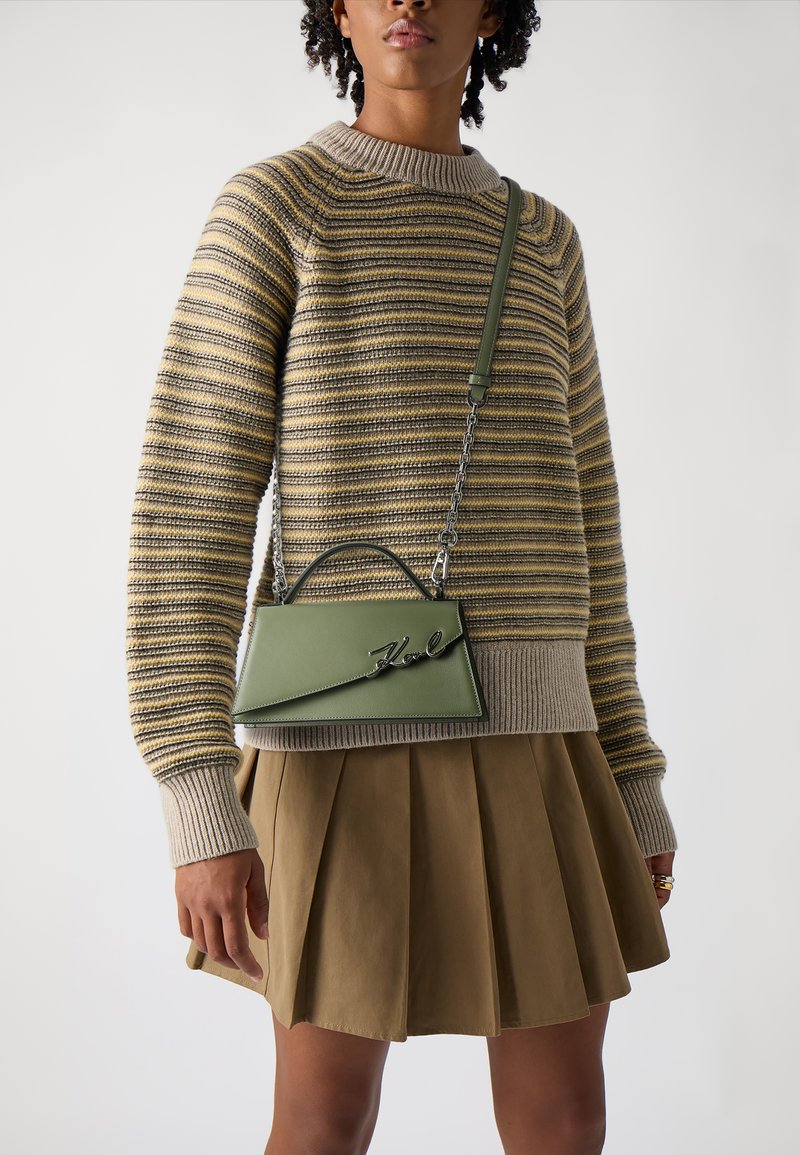 Bolsa de couro verde com uma forma estruturada, detalhe do logótipo em prata e uma alça de corrente, combinada com um suéter listrado de malha e uma saia plissada.