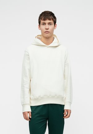 Jeune homme aux cheveux bruns courts portant un sweat à capuche blanc uni et un pantalon vert foncé, se tenant devant un fond clair.
