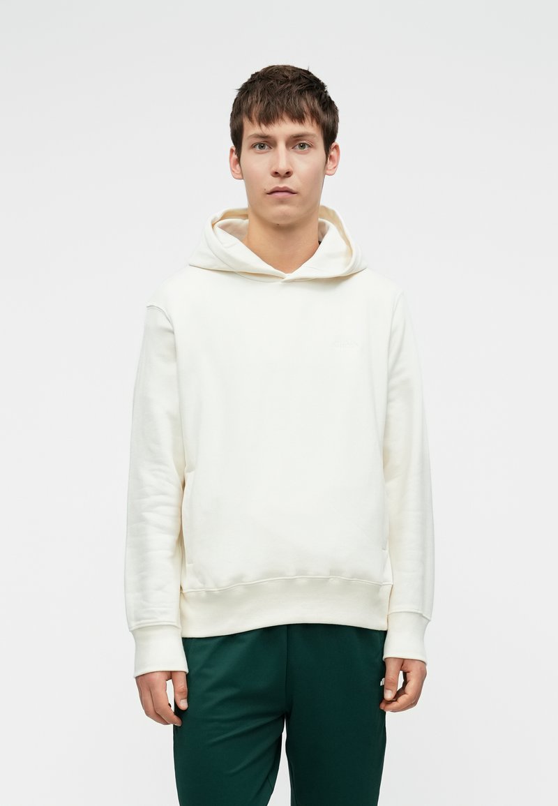 Jovem com cabelo castanho curto, vestindo um hoodie branco simples e calças verde-escuras, de pé contra um fundo claro.