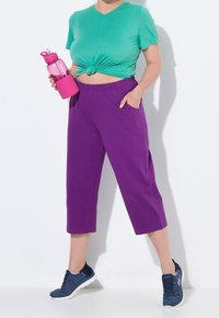 Groene korte mouwen knooptop, paarse cropped broek met zakken, en marineblauwe sportschoenen. Houdt een roze waterfles vast. Gladde texturen.