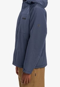 Granatowa kurtka z kapturem w kratę, z kieszenią na lewej piersi i logo marki na ramieniu, wykonana z lekkiego, wodoodpornego materiału.