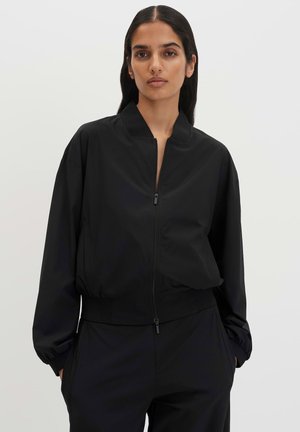 Frau mit langen dunklen Haaren, die eine schwarze Jacke mit Reißverschluss und eine passende schwarze Hose trägt, steht vor einem schlichten hellen Hintergrund.