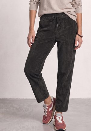 Personne portant un pantalon noir à cordon, un haut beige à manches longues et des baskets rouges et blanches debout sur un sol gris.