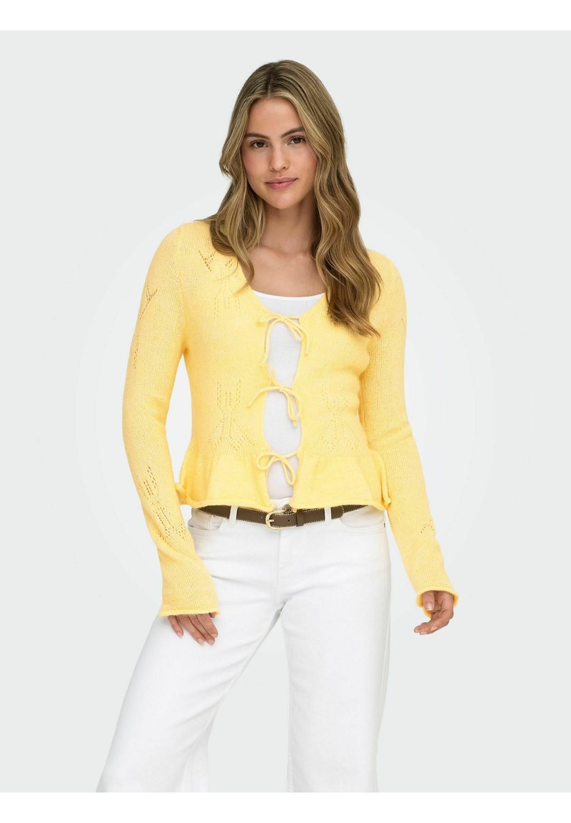 Yellow knit cardigan with a peplum hem, long sleeves, open front with lace-up detail, and star pattern accents. Worn over a white top.