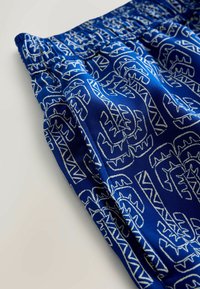 Rugalmas derékpántú, kék nadrág fehér absztrakt törzsi mintákkal, oldalzsebbel és részletes anyagtextúrával világos háttéren.