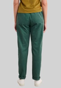 Grüne Baumwollhosen mit lockerer Passform, mit umgeschlagenen Säumen und Gesäßtaschen. Getragen mit weißen Sneakers und einem senfgelben Oberteil.