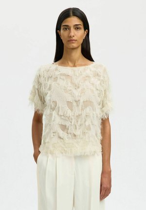 Kvinde med langt mørkt hår iført en tynd, struktureret kortærmet top og lyse, vidde bukser med høj talje, stående med hænderne i lommerne.