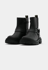Botas negras hasta el tobillo, hechas de cuero suave, con cremallera lateral, detalles de correas y una suela gruesa con dibujo para mayor tracción.