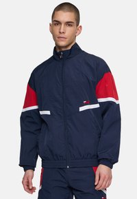 Giacca a vento navy con accenti rossi e bianchi, zip frontale, colletto alto e polsini elastici; caratterizzata da un logo del marchio sul petto.