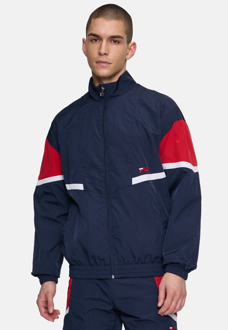 Giacca a vento navy con accenti rossi e bianchi, zip frontale, colletto alto e polsini elastici; caratterizzata da un logo del marchio sul petto.