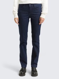 Pessoa vestindo jeans retos azul-escuro, camisa de mangas compridas branca e sapatos loafers pretos, posicionada contra um fundo claro e liso.