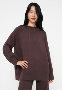 Mulher com cabelo preto longo, vestindo um capuz de malha solto em mauve escuro e calças combinando, de pé com uma mão na cintura.