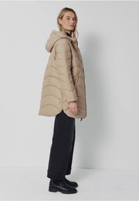 Beżowy ocieplany płaszcz z kapturem, charakteryzujący się pikowanym wzorem i bocznymi sznurkami. Noszony na czarnych spodniach i botkach za kostkę, prezentujący luźną sylwetkę.