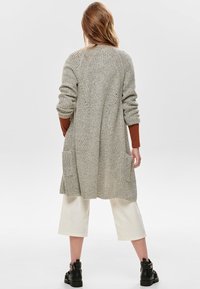 Lichtgrijze gebreide cardigan met een lange, open voorkant, geribbelde textuur, zijzakken en verlaagde schouders, gedragen over crèmekleurige wijde broekspijpen.