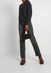 Pull noir en maille avec motif torsadé, jean en denim gris taille haute et bottines marron à talons. Tenant un petit sac à main noir tressé.