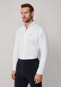 Chemise blanche à col pointu, ornée d'un petit logo brodé. Manches longues et coupe ajustée, associée à un pantalon sombre.
