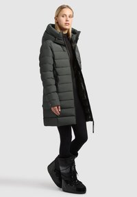 Tume roheline puffer-tüüpi mantel kõrge krae, tikitud tekstuuri, esiluku ja küljetaskutega, kombineeritud mustade talvesaabaste ja retuusidega.