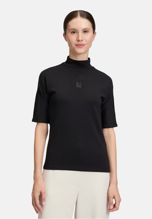Donna che indossa una maglietta nera a maniche corte con colletto alto e un piccolo logo sul petto, abbinata a pantaloni beige chiaro, in piedi su uno sfondo bianco.