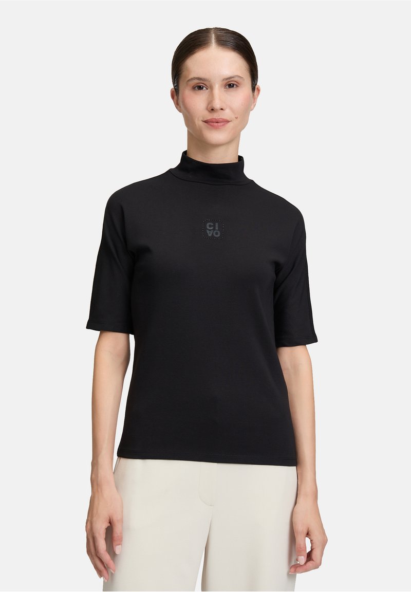 Femme portant un haut noir à manches courtes et col montant avec un petit logo sur la poitrine, et un pantalon beige clair, se tenant contre un fond blanc.