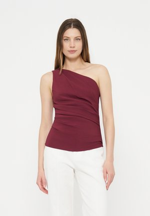 Femme portant un haut bordeaux sans manches à une épaule et un pantalon blanc, debout devant un fond blanc uni.