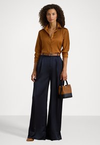 Καφέ σατέν πουκάμισο με κουμπιά, φαρδιά ναυτικά παντελόνια, δερμάτινη ζώνη με αγκράφα RL, κρατώντας μια μικρή καφέ και μαύρη τσάντα.