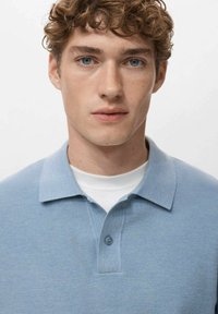 Jasnoniebieska koszulka polo z teksturowanym wzorem dzianiny, kołnierzyk zapinany na guziki, noszona na białej koszulce z okrągłym dekoltem. Gładki materiał, swobodny krój.