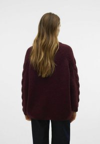 Bordeauxfarbener Pullover mit dickem strukturiertem Strick, ausgestattet mit geflochtenen Seitendetails, lockerer Passform und langen Ärmeln, kombiniert mit dunklen Hosen.