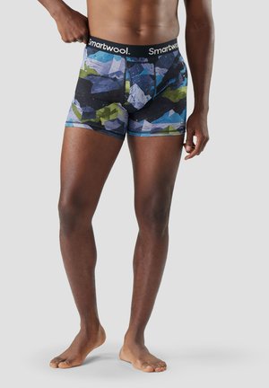 Hombre con calzoncillos tipo bóxer de patrones geométricos multicolores con cintura "Smartwool", de pie descalzo contra un fondo liso.