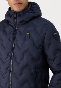 Giacca a vento blu navy con cappuccio, caratterizzata da un motivo a zigzag, zip frontale e toppe con logo sul petto e sulla manica.
