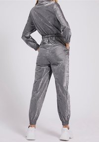 Mono metálico gris con cuello alto, cinturilla elástica y puños. Cuenta con bolsillos laterales y una superficie brillante y texturizada.