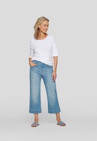 Weißes, strukturiertes Oberteil mit Wellenmuster, blaue weit geschnittene Jeans und blaue Sandalen. Leichte Stoffqualität und lockere Passform. Neutraler Hintergrund.