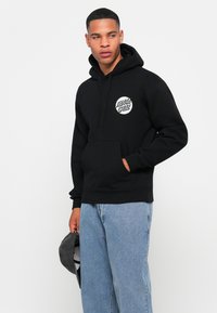 Moški nosi črno kapuco Santa Cruz in svetlo modre kavbojke, drži sivo kapo v levi roki in gleda desno.