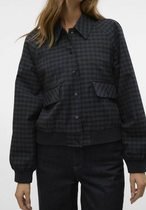 Woman wearing a dark checkered button-up jacket with flap pockets and ribbed cuffs, paired with dark denim jeans.