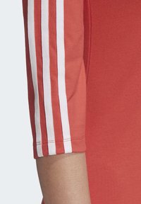 Rood sporttop met drie witte strepen langs de mouw, met een aansluitend ontwerp en zachte textuur. Schone afwerking aan de mouwzoom.