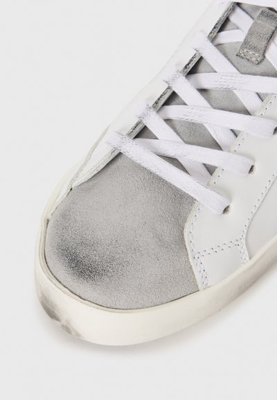 White sneaker with a gray suede toe cap, white laces, and a textured leather body. Features a rubber sole and stitching detail.